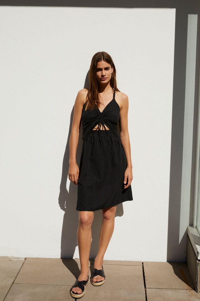 Else Ponza cotton ruched front cutout dress in black. Shown on model, front view. The dress features a v-neck ruched front with tie closure. The waistband has a cutout above the elastic. The skirt is an a-line shape that hits just above the knee. 