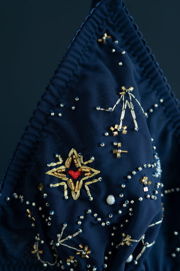 Close up detail view of the beading and embroidery on the Cygnus Bralette by Love & Swans. Deep blue mesh embroidered and beaded with constellation patterns in gold, silver, and red with pearls.