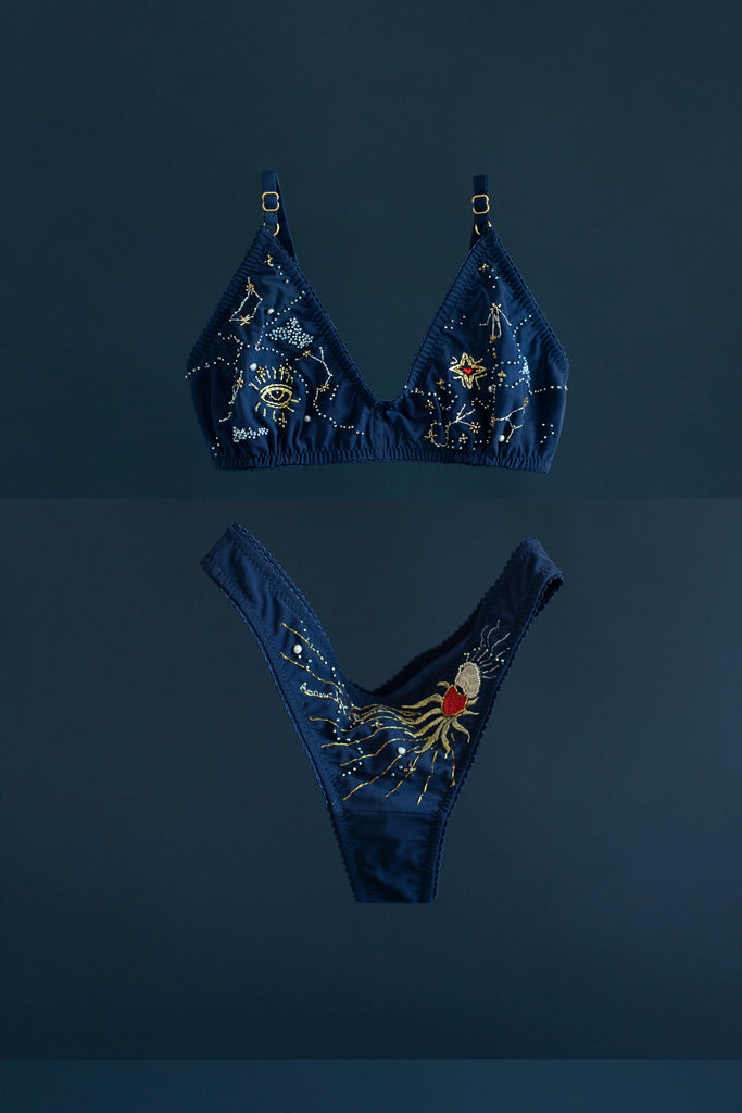 Cygnus Bralette and Eclipse Thong by Love & Swans, front view shown on dark teal background. The top features beaded & embroidered constellations on dark blue mesh. The thong features sun & moon beading and embroidery on dark blue mesh.