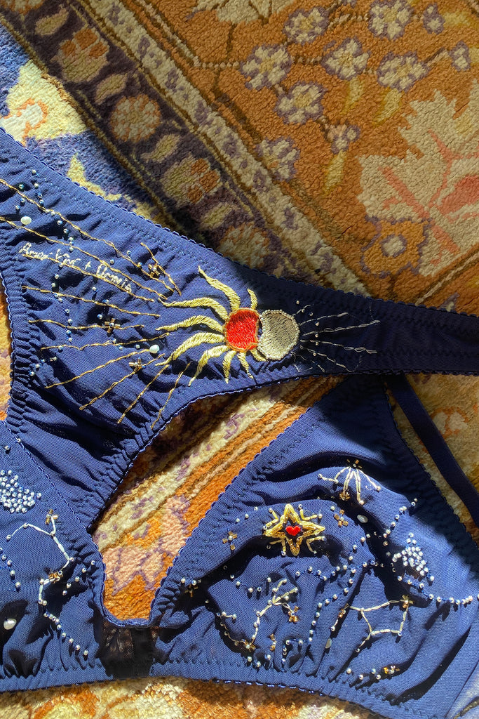 Close up view of Eclipse Thong by Love & Swans. Dark blue mesh is embroidered and beaded with a sun and moon crossing paths, rays emitting from both and the latin words for "love conquers all." Shown flat with the matching Cygnus bralette.