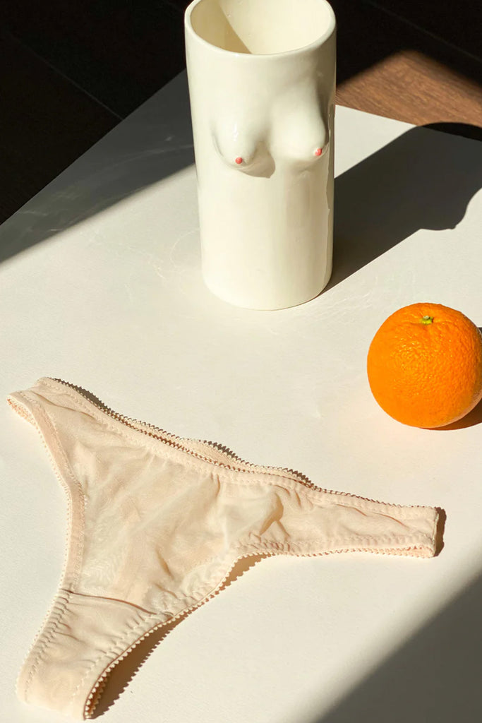 Love & Swans Easy Basic sheer mesh thong in cream beige. Shown flat on a table in a still life portrait wit ceramic boob vase and an orange. 