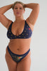 Eclipse Thong by Love & Swans. Dark blue mesh is embroidered and beaded with a sun and moon crossing paths, rays emitting from both and the latin words for "love conquers all." Front view on model. 