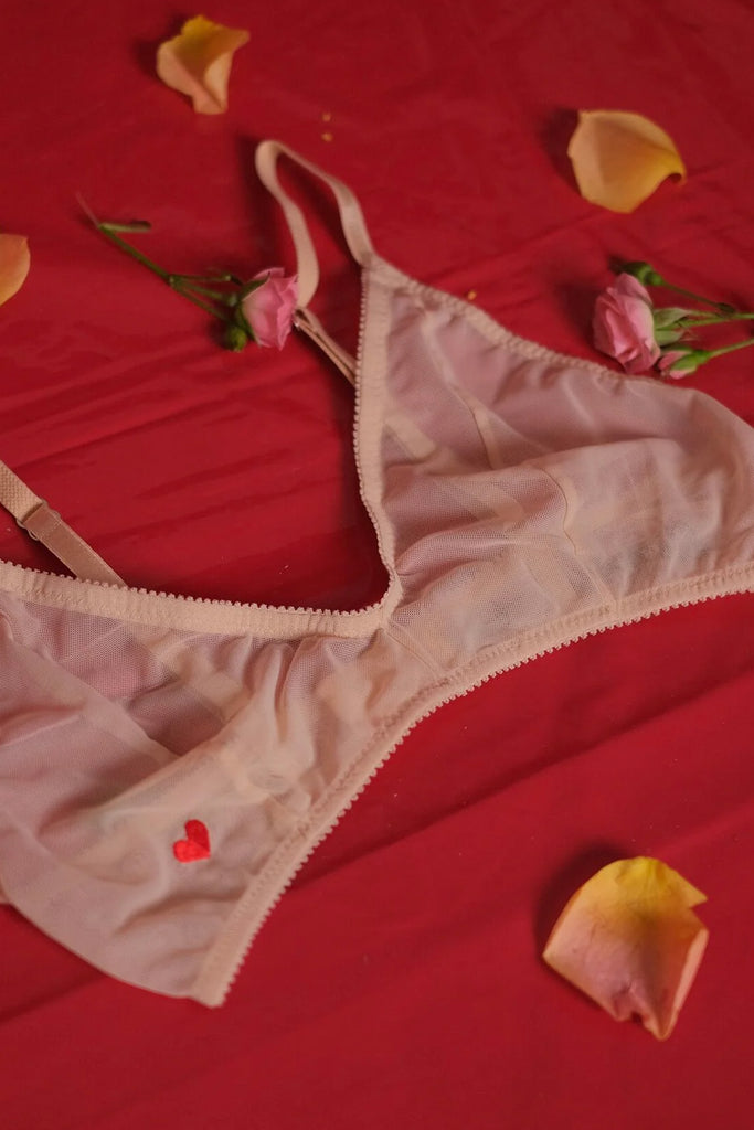 Love & Swans L'amour sheer beige mesh bralette, shown flat on red fabric surface surrounded by rose petals. Tiny heart embroidery is visible on bottom right cup.