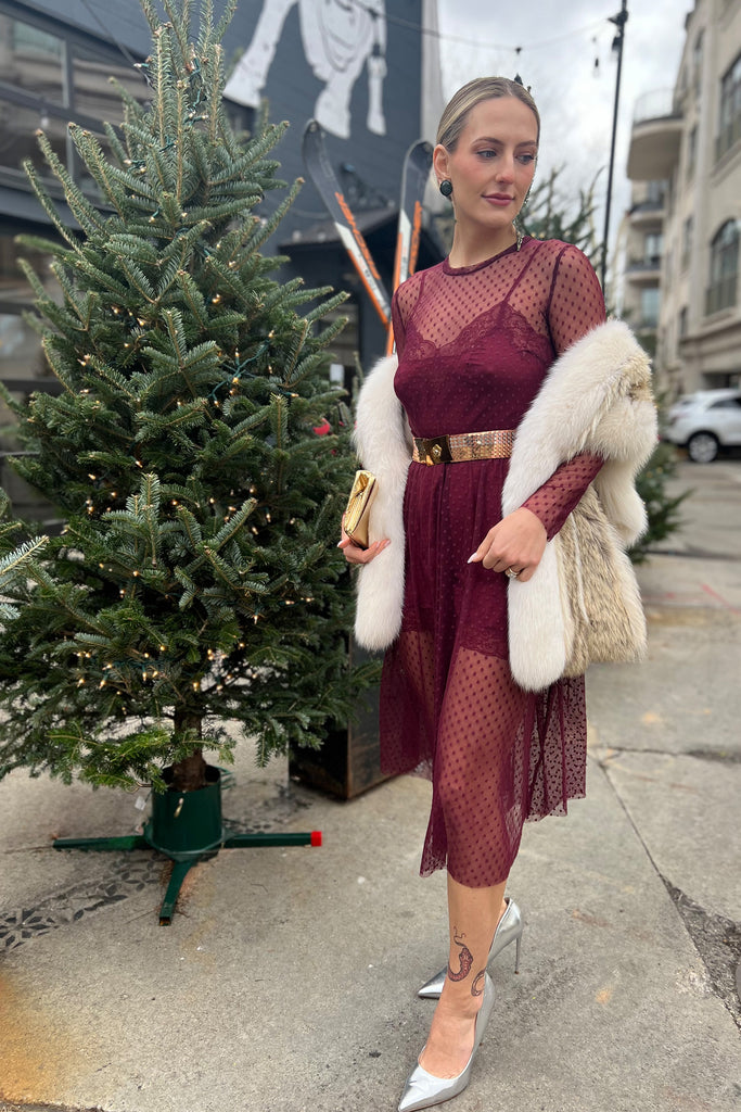 Only Hearts Silk Charmeuse Mini Slip in burgundy Dahlia, layered under the matching Coucou Lola long sleeve crew neck sheer polka dot mesh dress as a party outfit, styled on model with a fur vest, metallic belt, shoes, and clutch. 