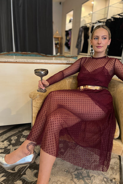 Only Hearts Coucou Lola Natasha Tea Dress in Dahlia burgundy sheer polka dot mesh, shown on model layered with the matching burgundy silk and lace slip dress and a metallic belt and shoes. Model is seated in a gold chair with champagne glass.