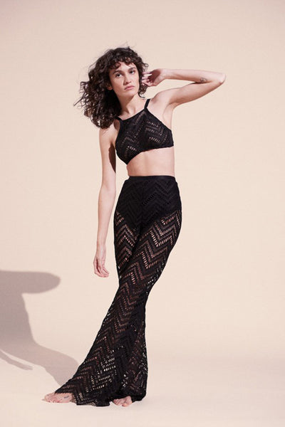 Paloma Casile Maxine black lace pants, shown on model, front view. Model is wearing matching lace halter style bralette. The pants have a black elastic waistband, built in opaque brief for modesty, and the sheer lace is a geometric chevron design with pointy angled hemline. 