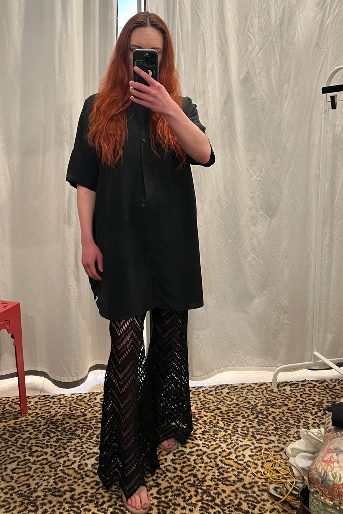 Paloma Casile Maxine black lace pants, shown on model, front view. Model is wearing a black oversized short sleeve button up shirt dress on top and taupe sandals. The pants have sheer lace in a geometric chevron design with pointy angled hemline. 