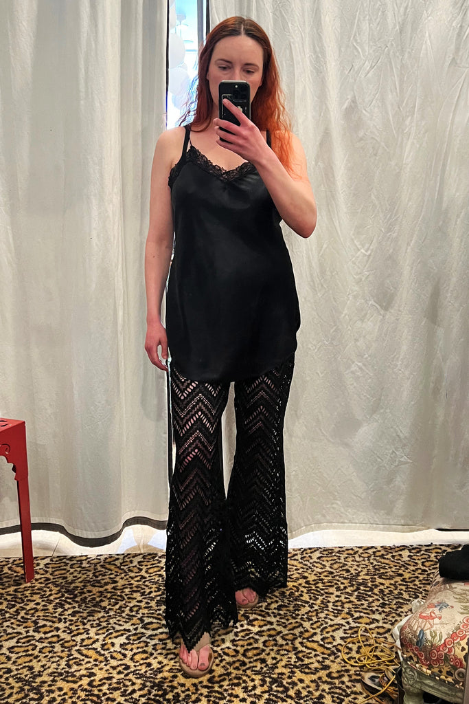 Paloma Casile Maxine black lace pants, shown on model, front view. Model is wearing a short black satin slip on top and taupe sandals. The pants have sheer lace in a geometric chevron design with pointy angled hemline. 