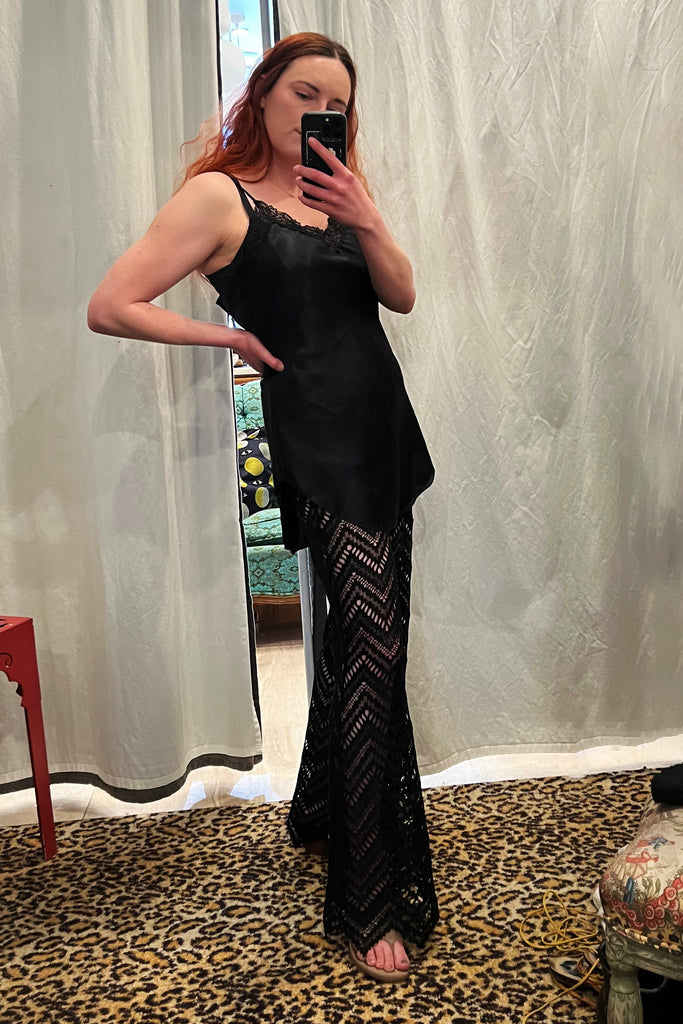 Paloma Casile Maxine black lace pants, shown on model, front/side view. Model is wearing a short black satin slip on top and taupe sandals. The pants have sheer lace in a geometric chevron design with pointy angled hemline.