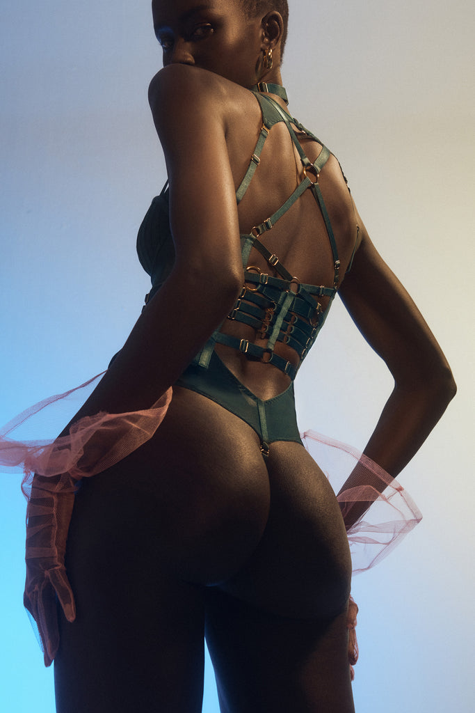 Bordelle Kora Basque in rich Eden green. Shown on model, back view, layered over the matching Kora Eden green sheer mesh bodysuit. Basque features many narrow satin elastic straps across the mid back and making an X shape across the shoulder with a gold plated ring in the center. .