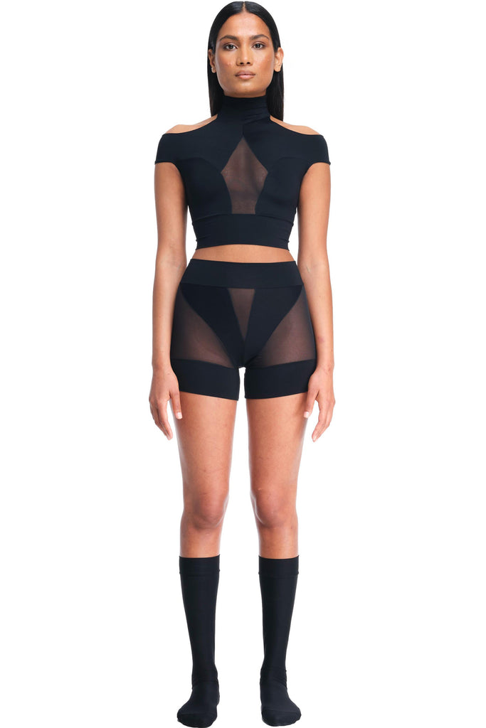 DSTM Innsai Brief in black, shown on model with the matching top. Shorts feature opaque panels with sheer inserts at the mid front and side hip/legs. The briefs are like shorts, with the hemline on the upper thigh. Front view.