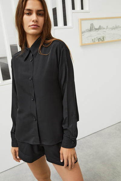 Else Diana silk shirt and shorts set in black. Shown on model, front view. The shirt is buttoned up, has a collar with pleated detail and long sleeves with pleated cuffs. The shorts have the same pleated detailing at the hemline.