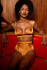 Amber orange Petra soft cup bralette by Studio Pia. Front view on model with matching high waist thong, choker, and garters. The model is seated in a relaxed manner on a red upholstered booth.