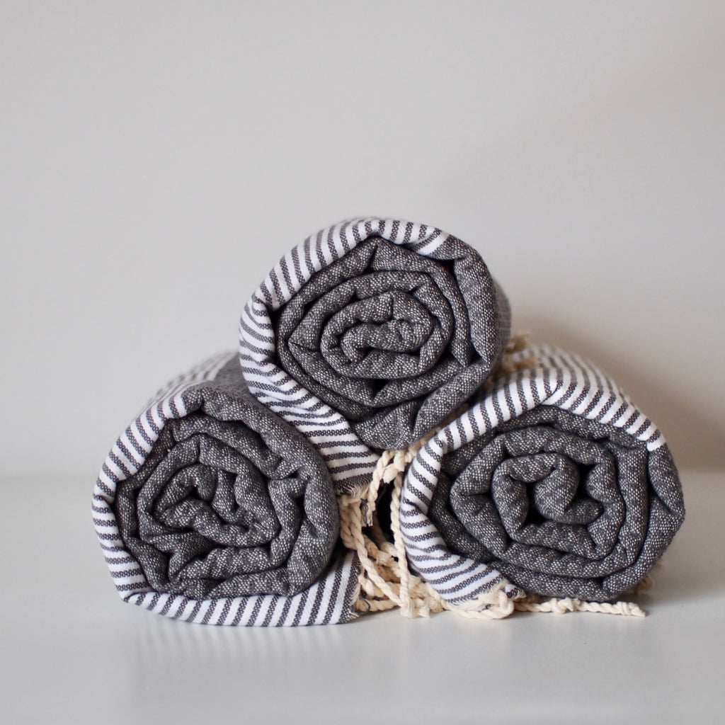 Three gray cotton hammam towels with fringed ends, rolled up and stacked in a pyramid arrangement. They are a dark gray with gray and white striped section and a waffle texture section.