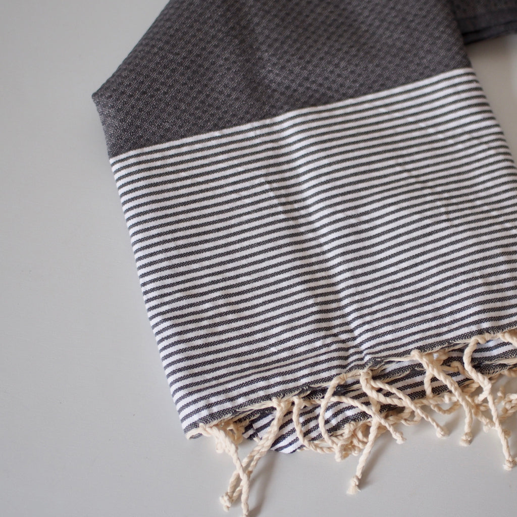 Gray cotton hammam towel, shown flat to show the braided fringe ends, white and gray striped section, and waffle texture dark gray section.
