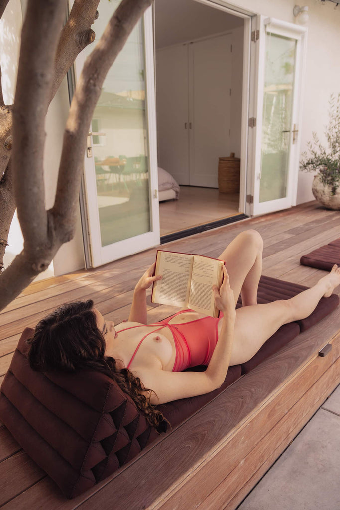 Model lounges on the deck while reading a book, wearing sheer cupless coral reversible la fille d'o bodysuit with open stomach.