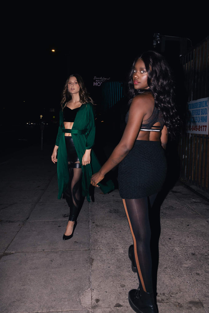 Two models walking at night on an urban sidewalk next to a fence. Right model wearing tight mini skirt with black Swedish stockings with brown stripe up the side.