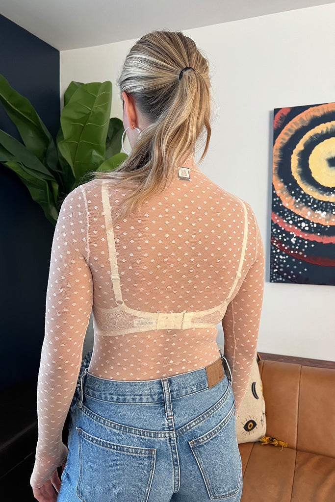 Only Hearts Coucou Lola long sleeve turtleneck bodysuit in light pink/beige tinted, back view shown on model. Image shows bodysuit with ivory lace bralette underneath, polka dot mesh, and styled with light wash jeans and silver jewelry. 